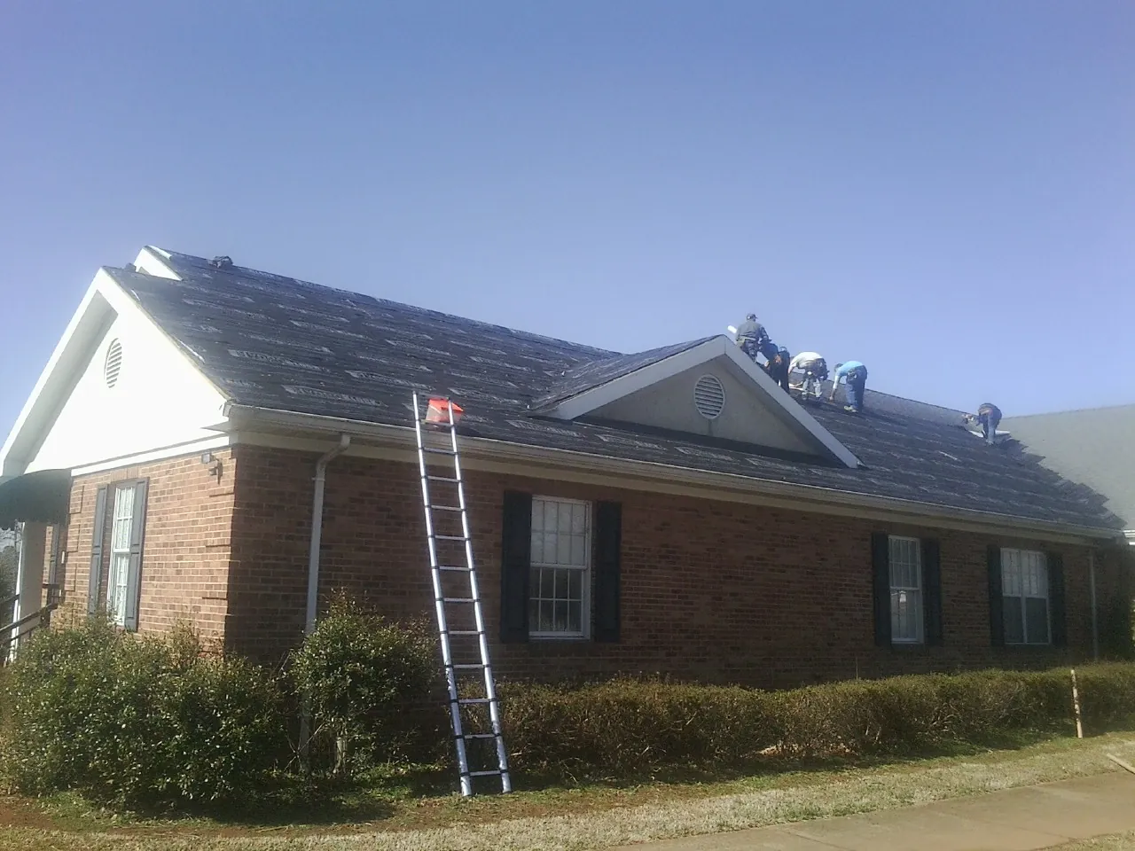 Crew tearing shingles off a brick ranch roof with a ladder set — Emergency Roof Repair work by our crew in Delmont