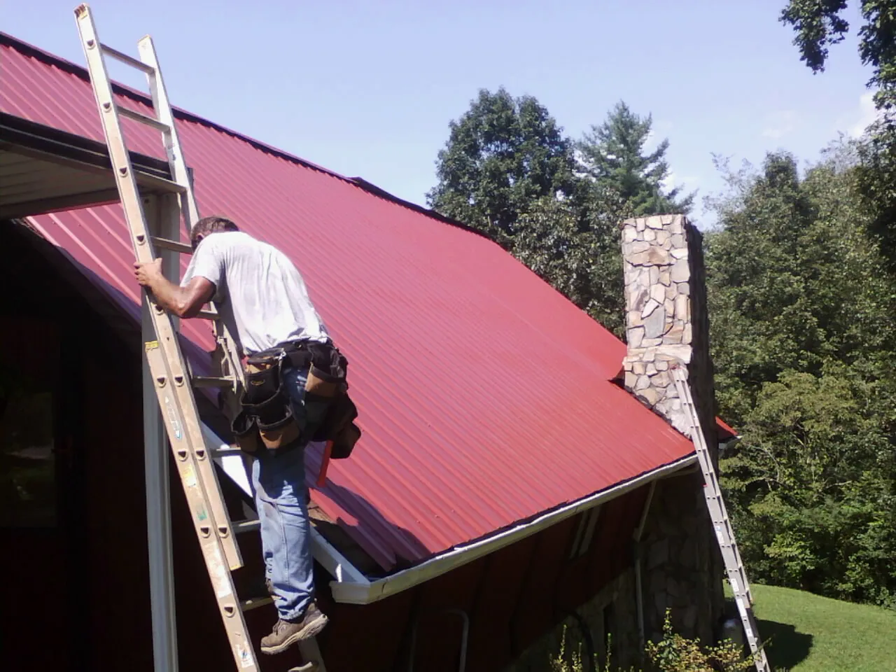 Roofer on a ladder beside a red metal roof and stone chimney — Soffit & Fascia Repair by Roofing Brothers in Delmont